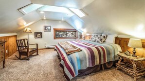 Un dormitorio con techo inclinado, tragaluces, una cama con colcha colorida, una silla de madera, una mesita de noche con lámpara y cuadros enmarcados en la pared.