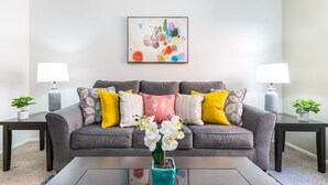 Ein modernes Wohnzimmer mit einem grauen Sofa, gelb-weißen Kissen, einer petrolfarbenen Vase mit weißen Blumen und abstrakter Kunst an der Wand.