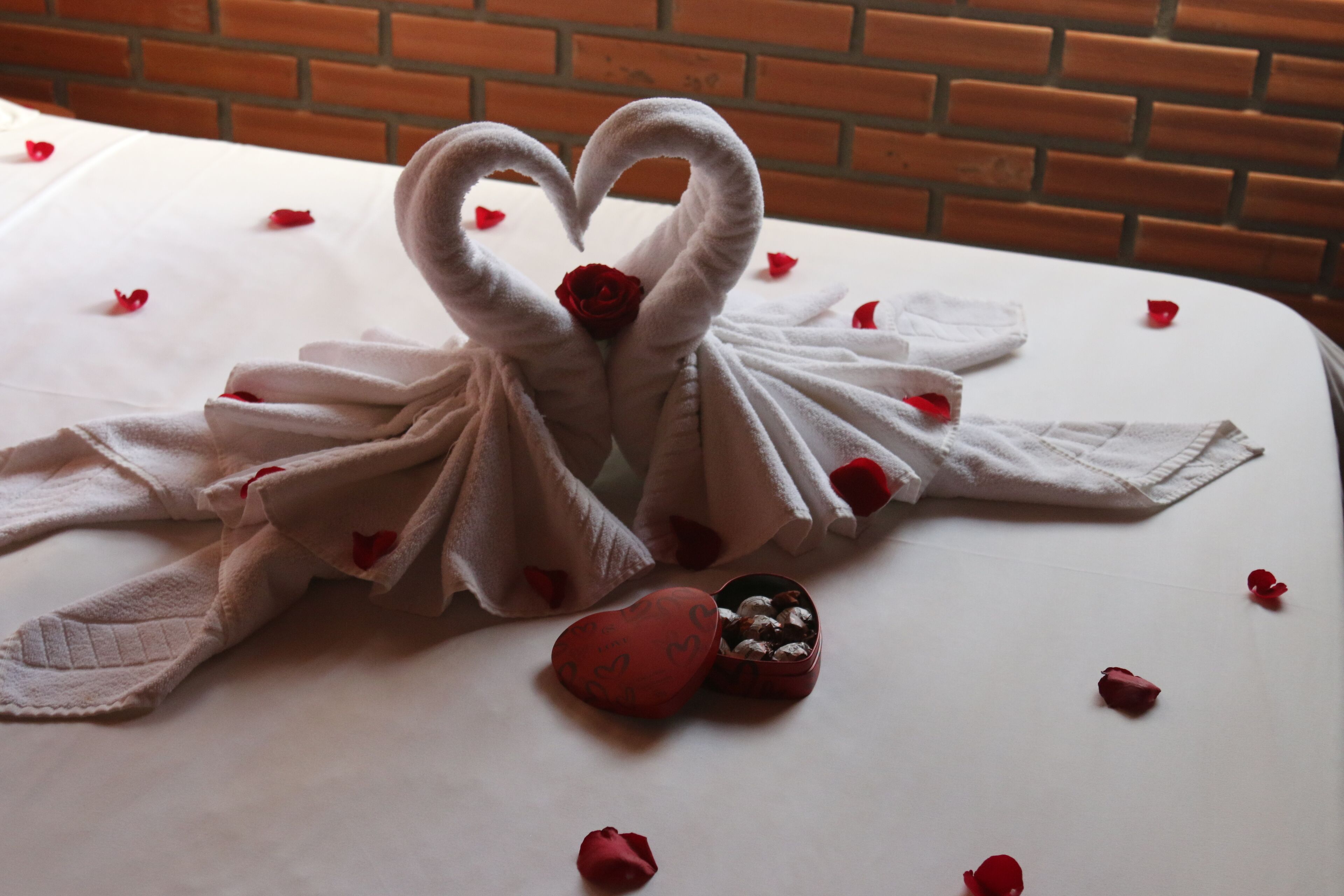 Una cama con una toalla dispuesta en forma de corazón y un pequeño objeto rojo con forma de corazón sobre la colcha.