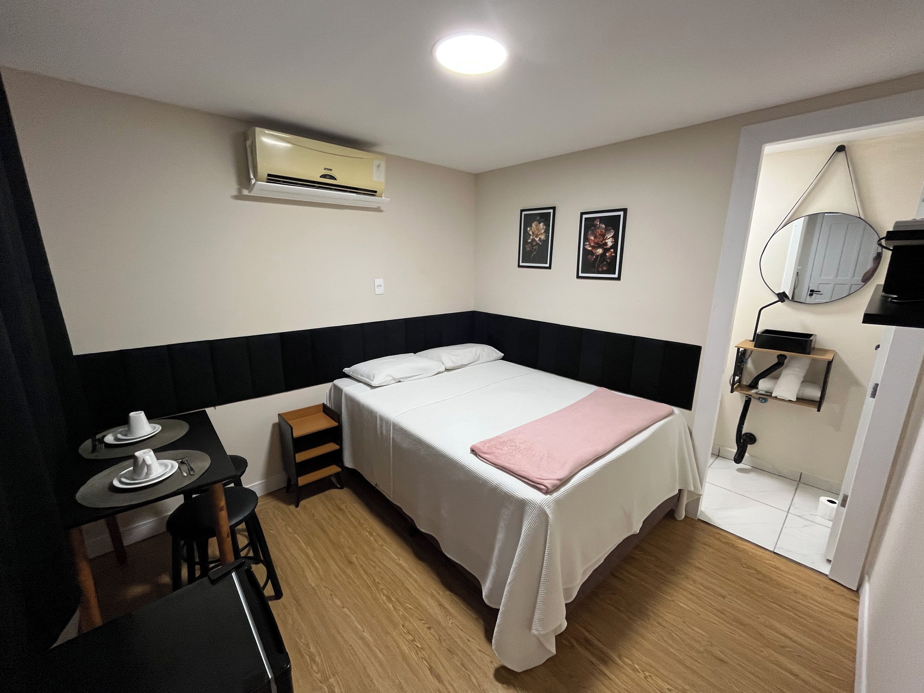 A hotel room with a bed, a desk with a chair, an air conditioning unit, a small shelf with towels, and a mirror on the wall.