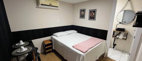 A hotel room with a bed, a desk with a chair, an air conditioning unit, a small shelf with towels, and a mirror on the wall.
