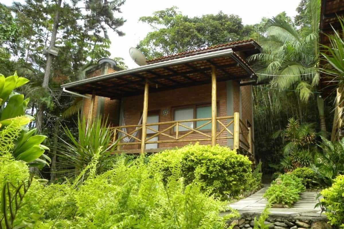 A rustic wooden house with a balcony, surrounded by lush greenery and tropical plants.