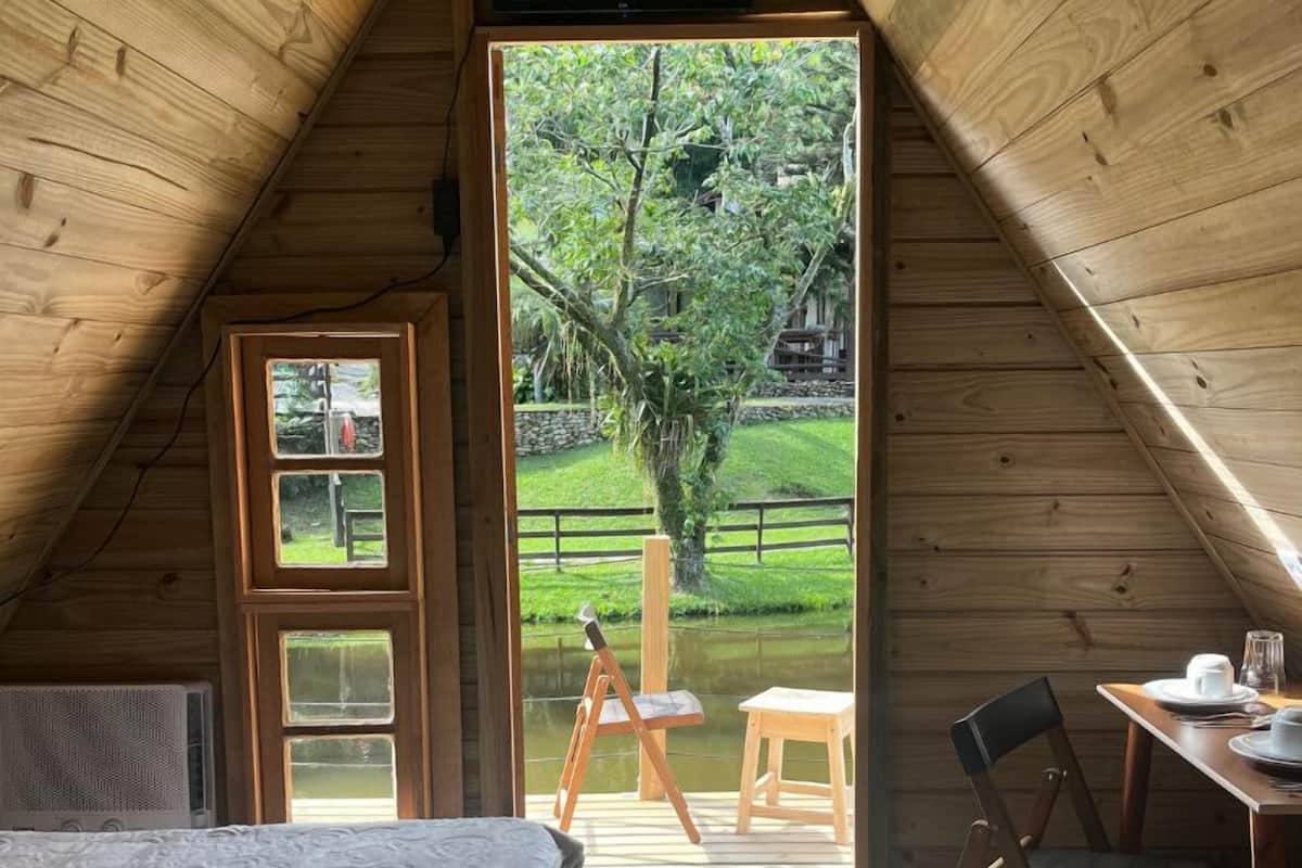 A cozy attic bedroom with a wooden sloped ceiling, a bed with a white comforter, a small table with a chair, and a view of a garden through a glass door.