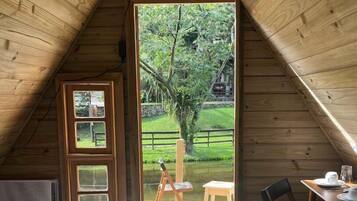 A cozy attic bedroom with a wooden sloped ceiling, a bed with a white comforter, a small table with a chair, and a view of a garden through a glass door.