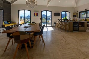 Un vaste espace de salle à manger avec une grande table en bois et des chaises modernes, une cuisine dotée d’un îlot central et de grandes fenêtres offrant une vue sur les montagnes.