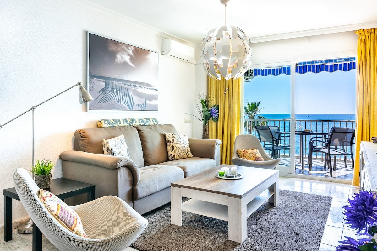 A living room with a sofa, armchairs, a coffee table, and a view of the sea through a balcony.
