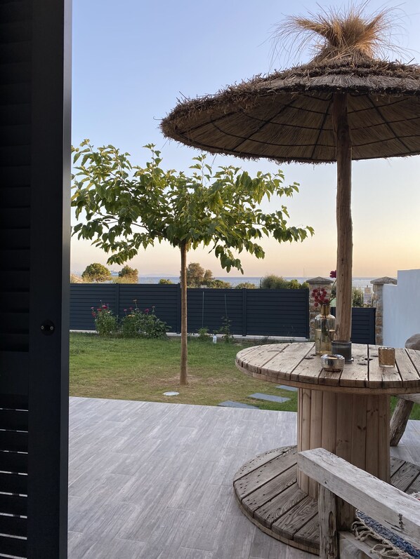 Une terrasse en bois avec une table ronde et un parasol en chaume, donnant sur une cour verdoyante avec un arbre et une clôture.