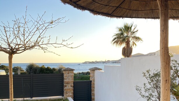 Une terrasse avec une table en bois et un parasol en chaume, un mur de pierre et une vue sur la mer.