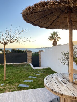 Une terrasse avec une table en bois et un parasol en chaume, un mur de pierre et une vue sur la mer.