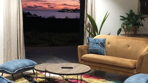 Un salon comprenant un canapé, deux fauteuils, une table basse et un tapis coloré. Une grande fenêtre, ornée de rideaux, offre une vue sur la mer et sur le coucher de soleil.