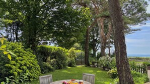 Une table ronde en bois, entourée de chaises, un bouquet de fleurs, une assiette de fruits et un verre de jus d’orange.