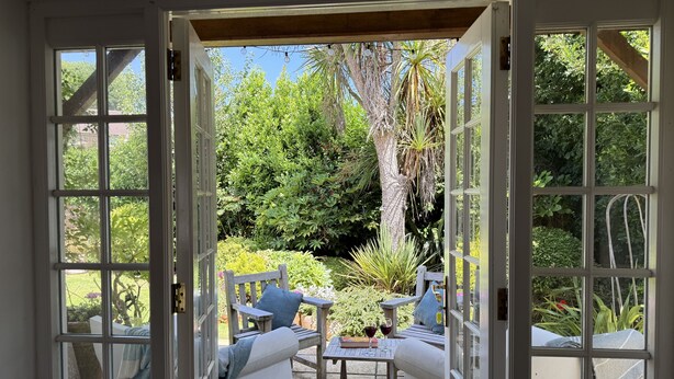 Une terrasse ouverte avec deux chaises en bois et une table, un canapé blanc et une vue sur un jardin luxuriant, visible à travers des portes ouvertes.