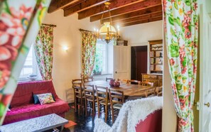 A cozy room with a wooden ceiling, a dining table with chairs, a sofa with a cushion, and a bookshelf.