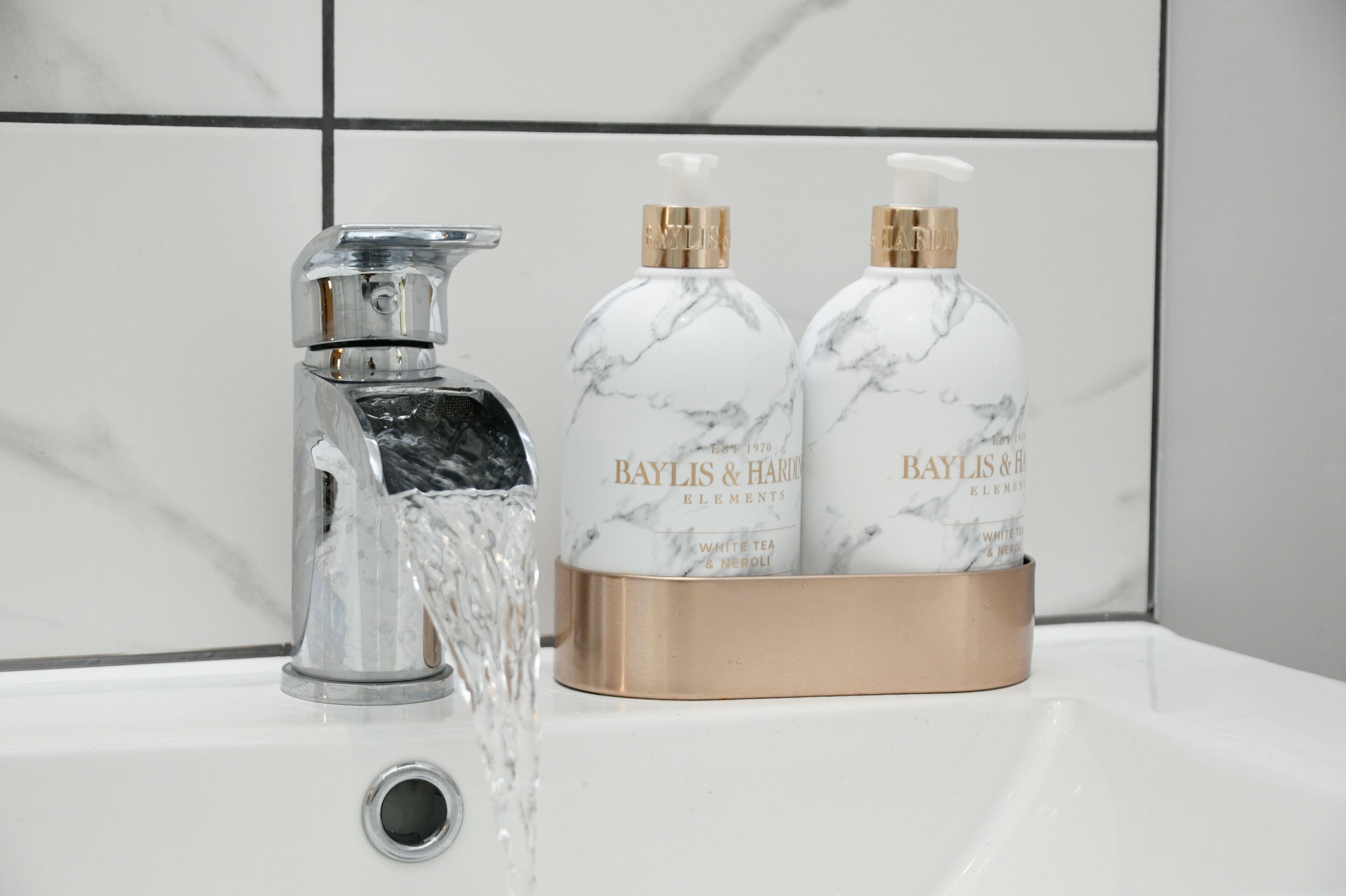 A bathroom sink with a marble faucet and two bottles of Baylis & Harrold Elements white tea and neroli.
