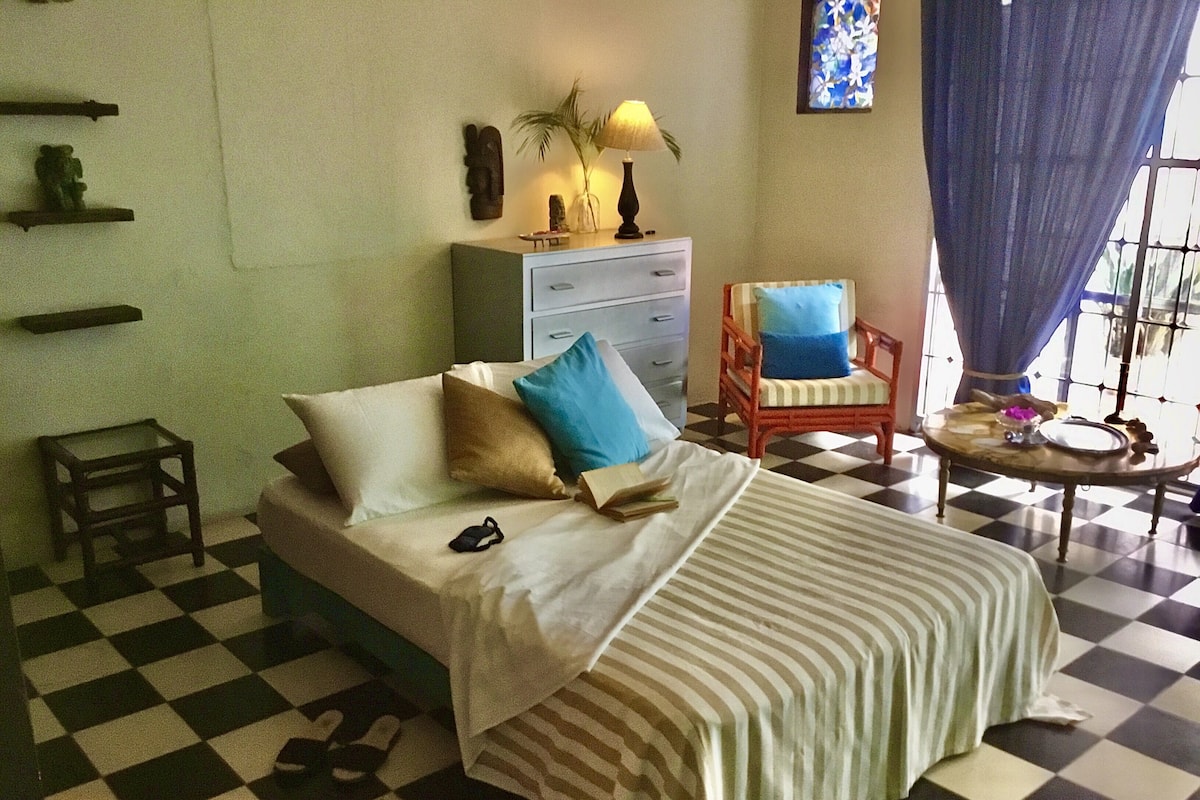 Ein Schachbrettmuster bedeckter Boden, ein Bett mit Kissen, ein Sessel, ein kleiner Tisch mit einer Lampe und eine Wand mit einem Fenster und Vorhängen.