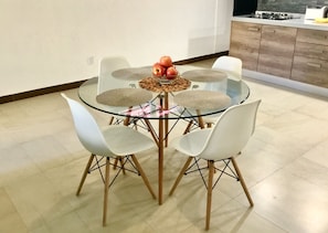 A modern dining area with a glass table surrounded by white chairs, a bowl of apples, and a woven placemat.