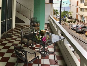 A balcony with a checkered floor, a glass table with chairs, and a view of the street and ocean.