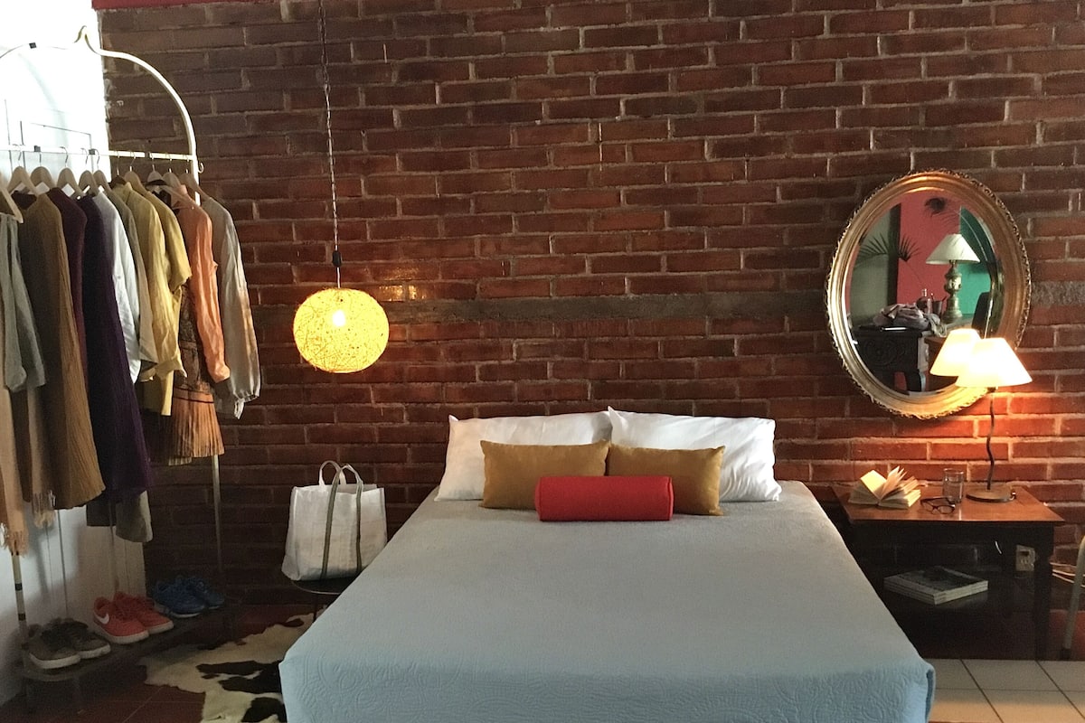 Ein Hotelzimmer mit Backsteinwand, einem Bett mit weißer und brauner Bettwäsche, einer Pendelleuchte, einem Spiegel und einem Nachttisch mit Lampe und Büchern.