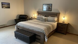 Un dormitorio con una cama grande, una silla, un banco, dos mesitas de noche con lámparas y un cuadro en la pared.