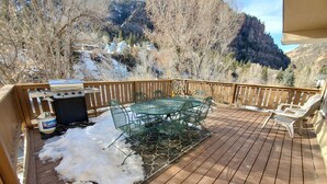 Una terraza de madera con una mesa y sillas de metal, rodeada de nieve y árboles.