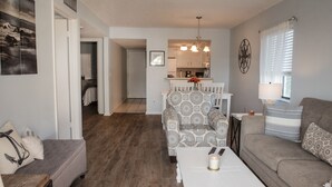 A living room with a sofa, a patterned armchair, a coffee table, and a dining table with chairs. There is a kitchen in the background with a window and a lamp. - Beautiful Condo steps away from Beach (St. Augustine Beach)