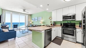 Cocina moderna con armarios blancos, microondas de acero inoxidable y horno. Zona de comedor con sofá azul y vista al océano a través de amplios ventanales.