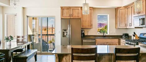 A modern kitchen with wooden cabinets, a central island with bar stools, stainless steel appliances, and a view of the outdoors through large sliding glass doors.