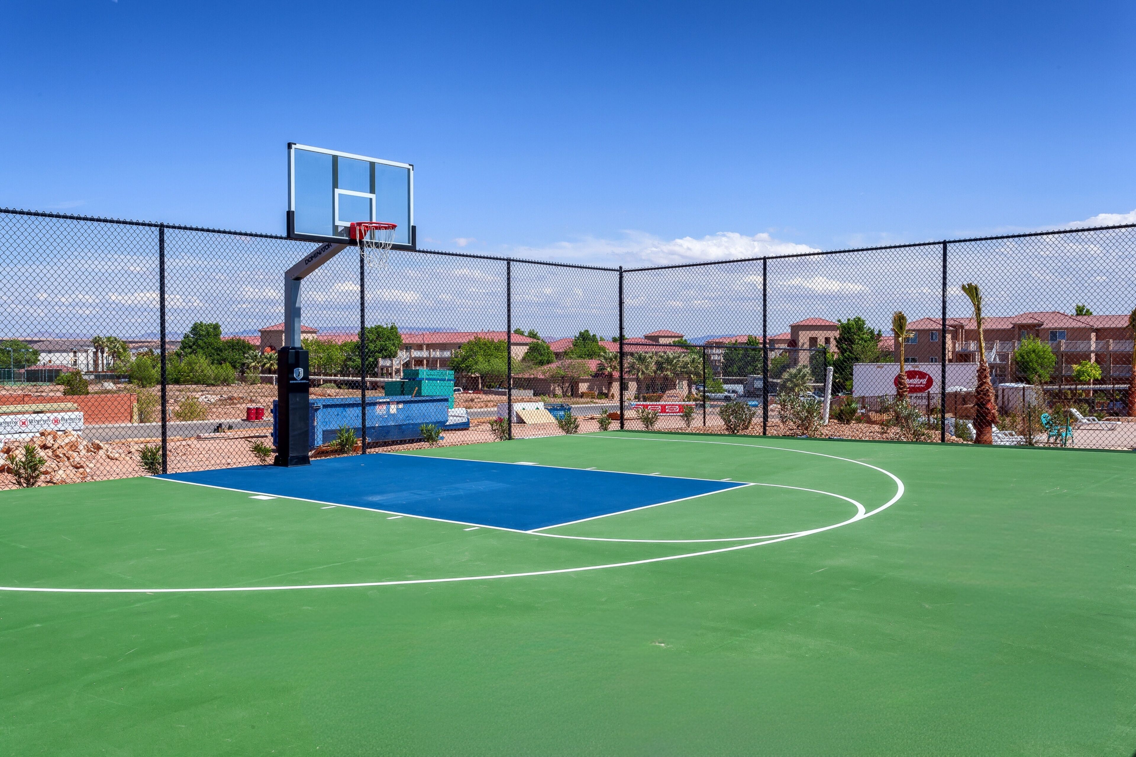 An outdoor basketball court with a chain-link fence, a blue playing surface, and a basketball hoop.