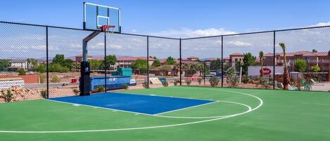 An outdoor basketball court with a chain-link fence, a blue playing surface, and a basketball hoop.