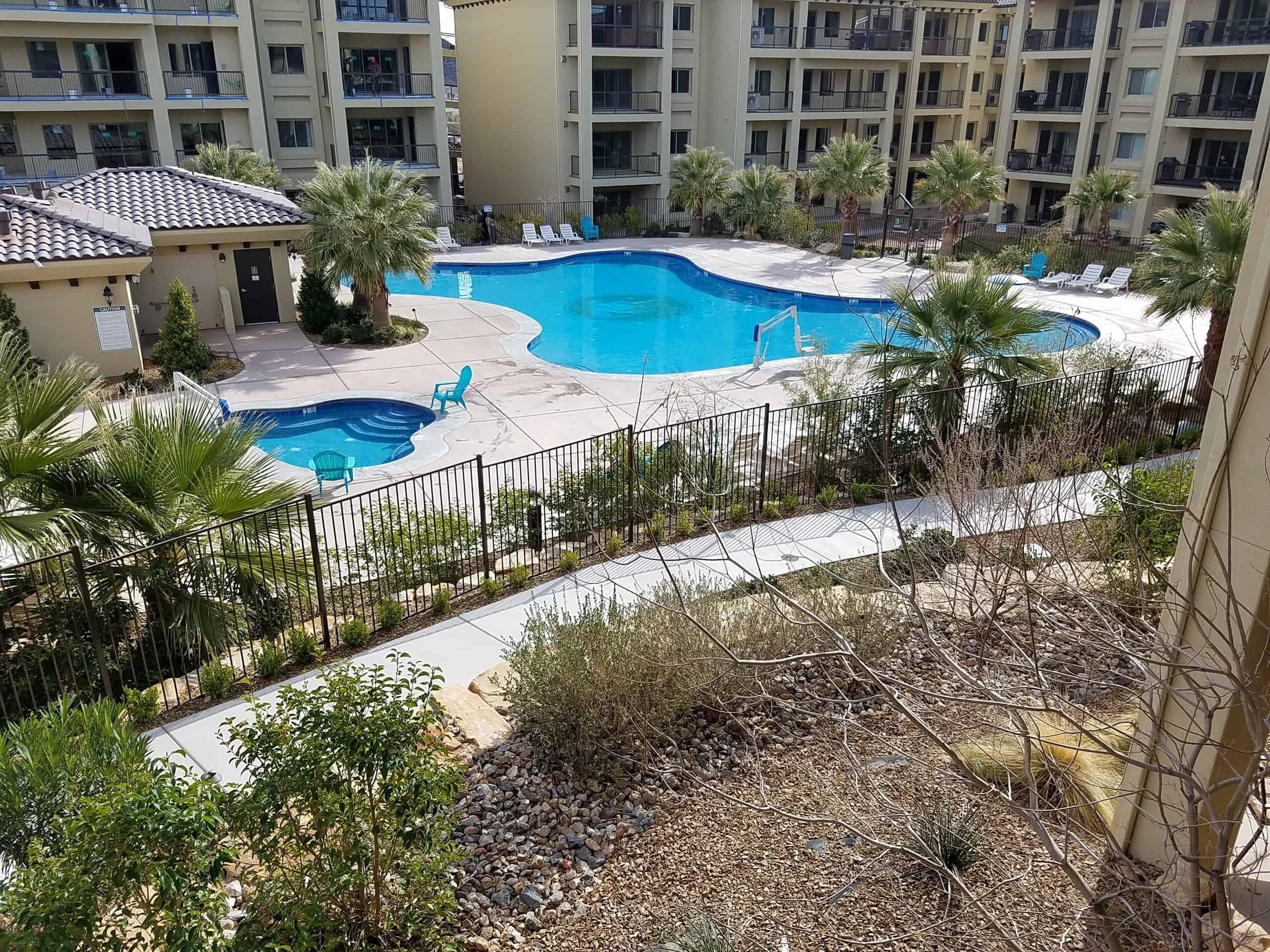 A swimming pool surrounded by lounge chairs and palm trees, with a multi-story building in the background.