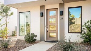 A modern building entrance with a glass door, a number '24' sign, and a small garden area with plants and gravel.