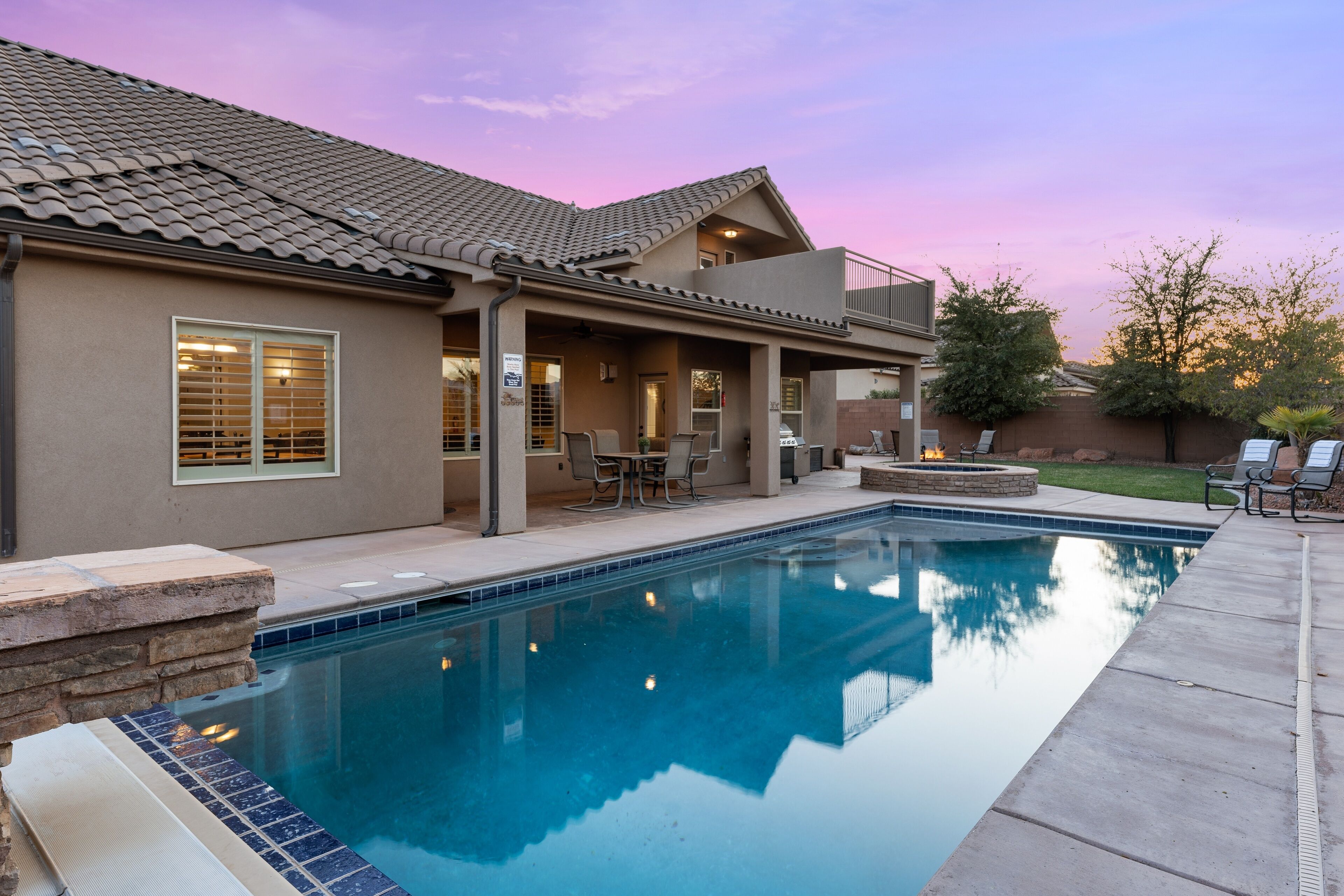 A residential pool area with a clear view of the pool, patio furniture, and a two-story house with a tiled roof.