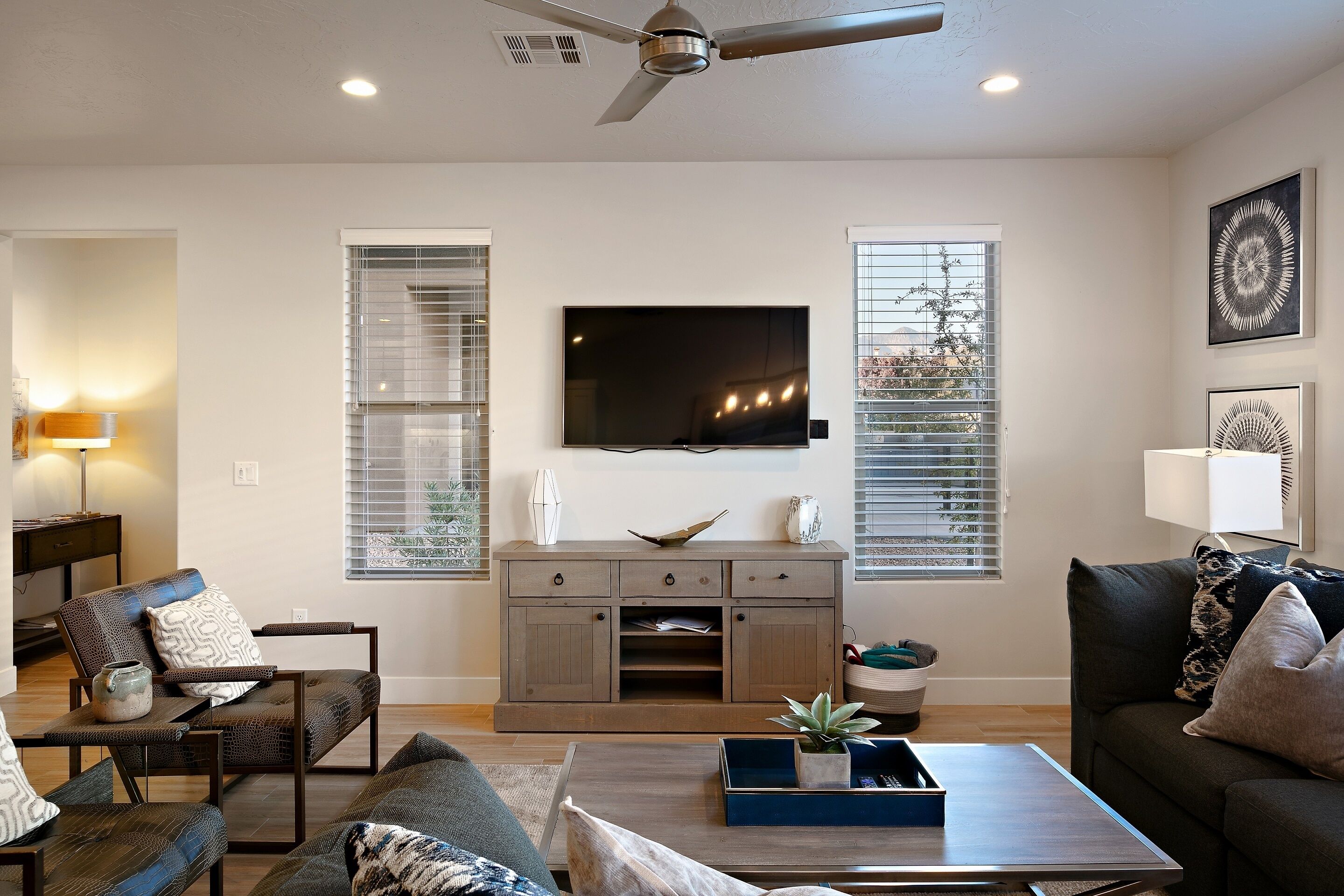 A modern living room with a flat-screen TV, a ceiling fan, and a large window with blinds.