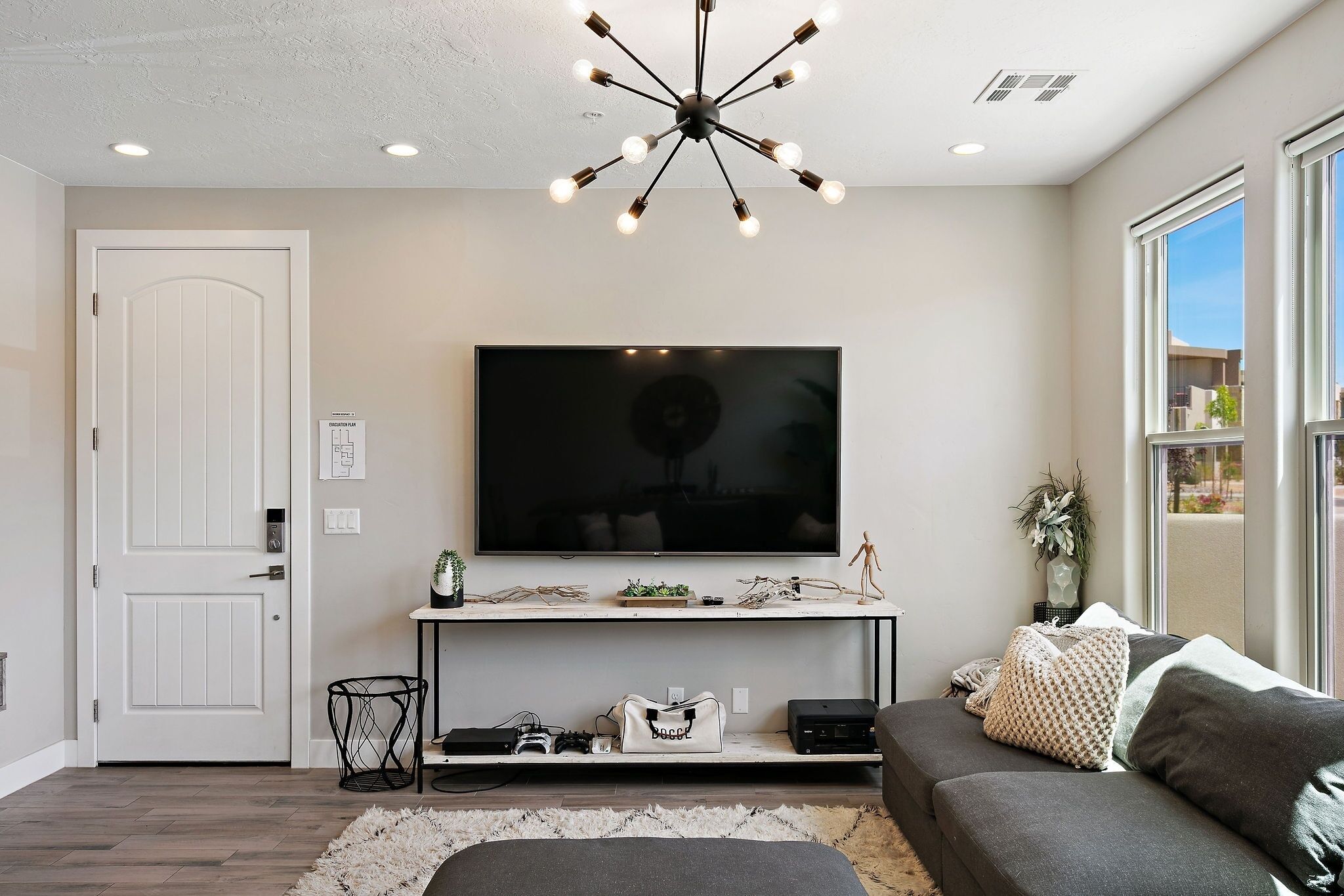 A modern living room with a flat-screen TV, a console table with decorative items, a grey sofa, and a large window providing natural light.
