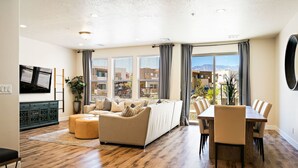 A modern living room with a sofa, ottomans, a dining table with chairs, and a flat-screen TV mounted on the wall. - Fun Times & Tan Lines by RedAwning (Washington)