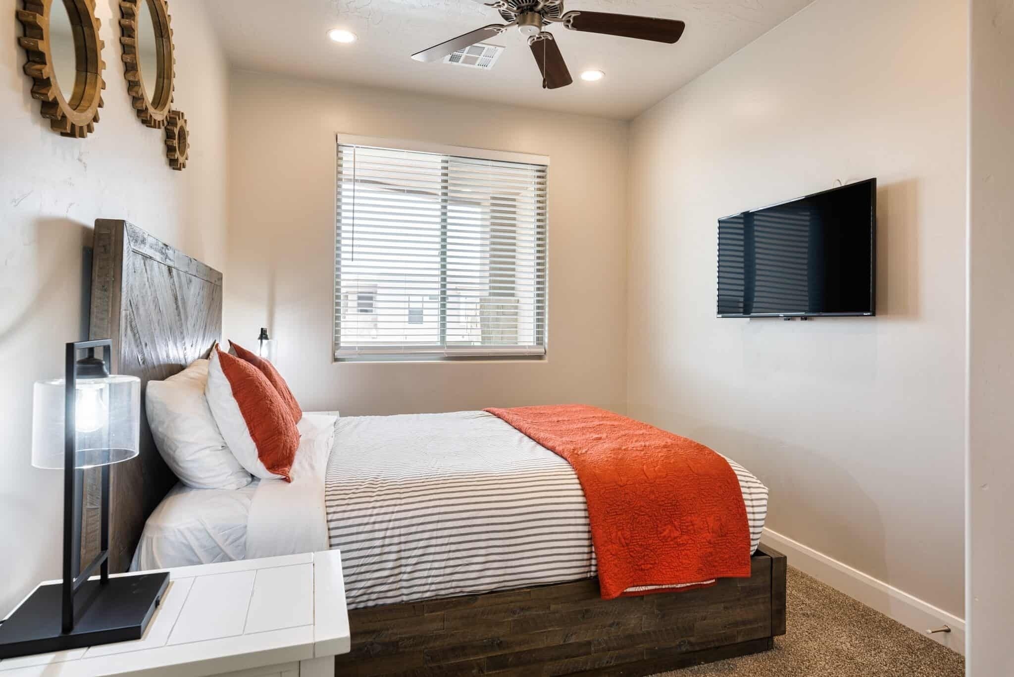 A bedroom with a bed, a desk, a ceiling fan, a window with blinds, and a TV mounted on the wall.