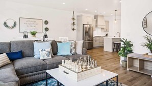 Una sala de estar moderna con un tablero de ajedrez como centro, un sofá gris con cojines azules, y una cocina al fondo.