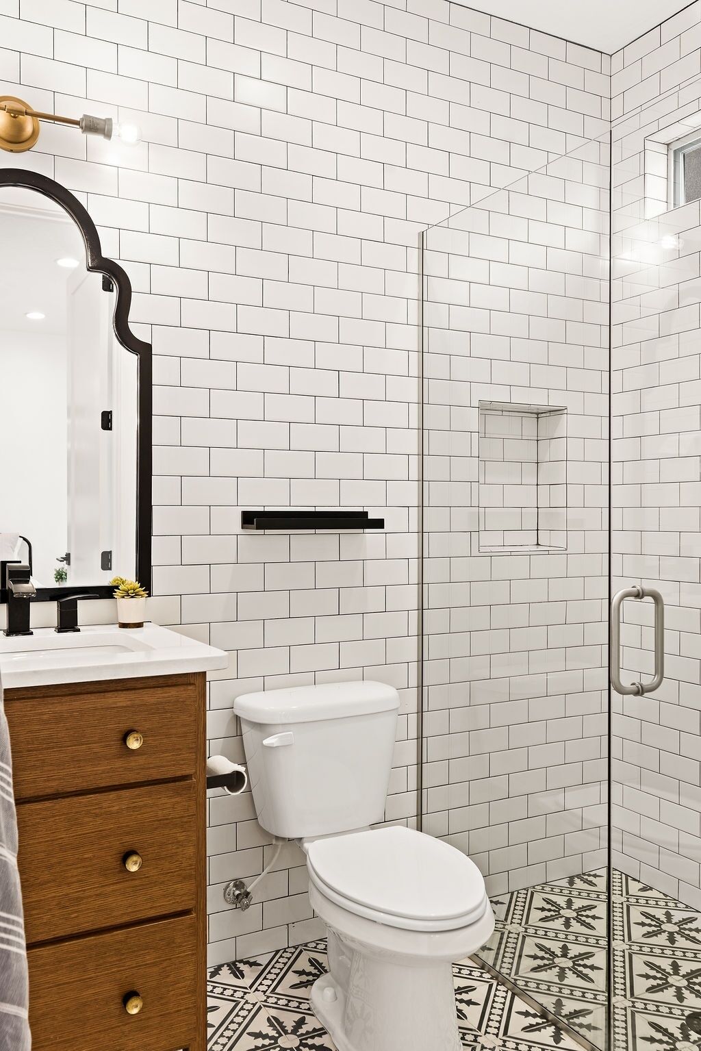 A bathroom with white subway tiles, a wooden vanity with brass handles, a white toilet, and a glass shower enclosure.