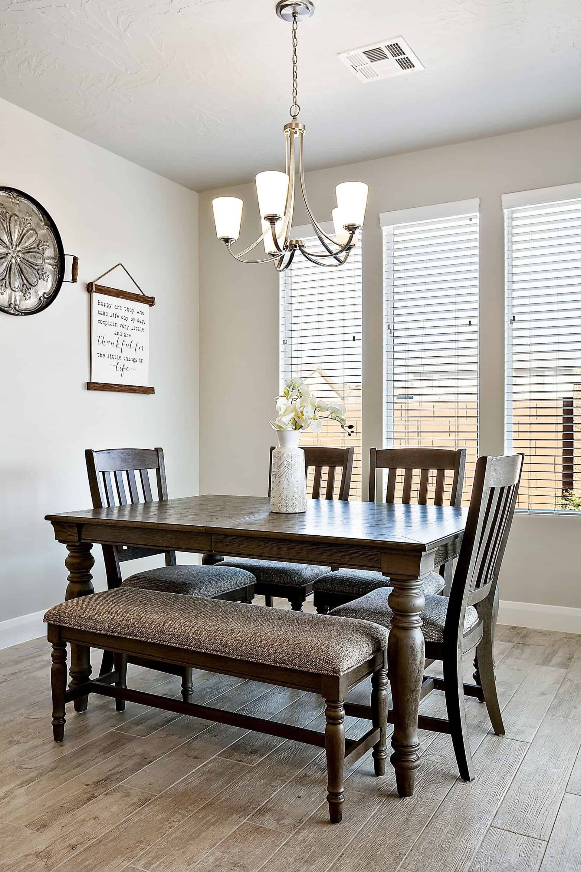 A dining room with a wooden table, four chairs, a bench, a chandelier, and a window with blinds.