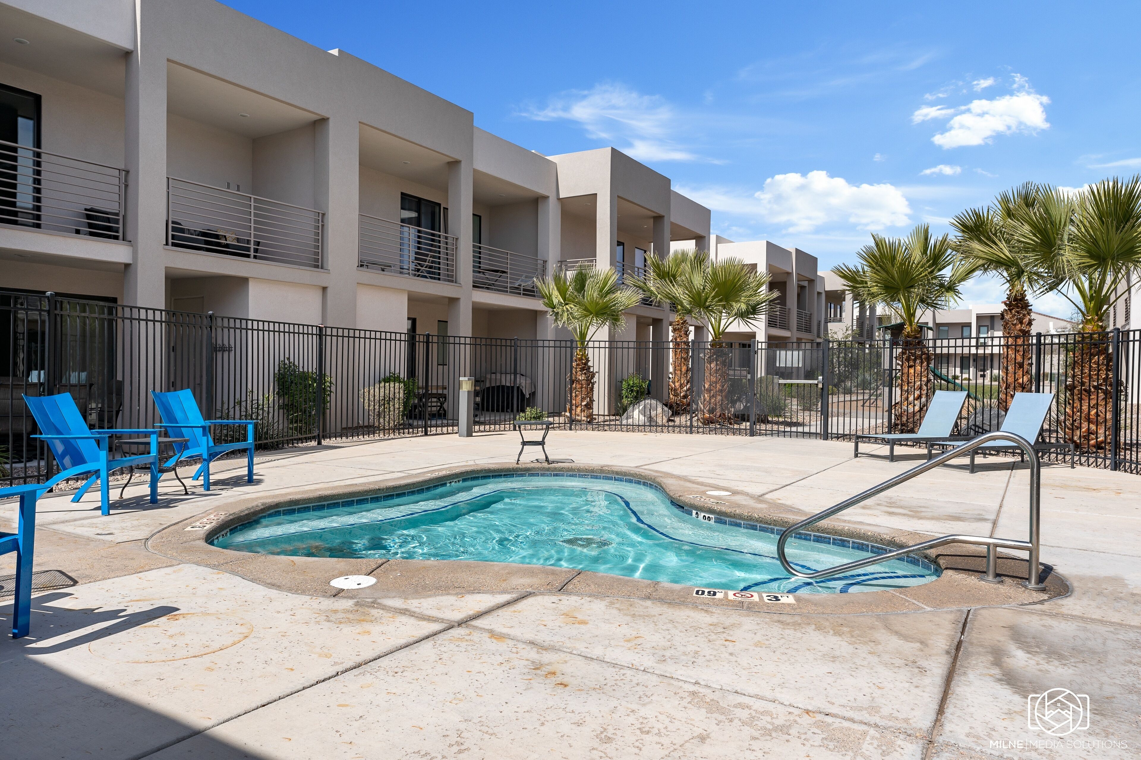 A swimming pool with blue tiles, surrounded by a concrete deck, and adjacent to a multi-story building with balconies and palm trees.