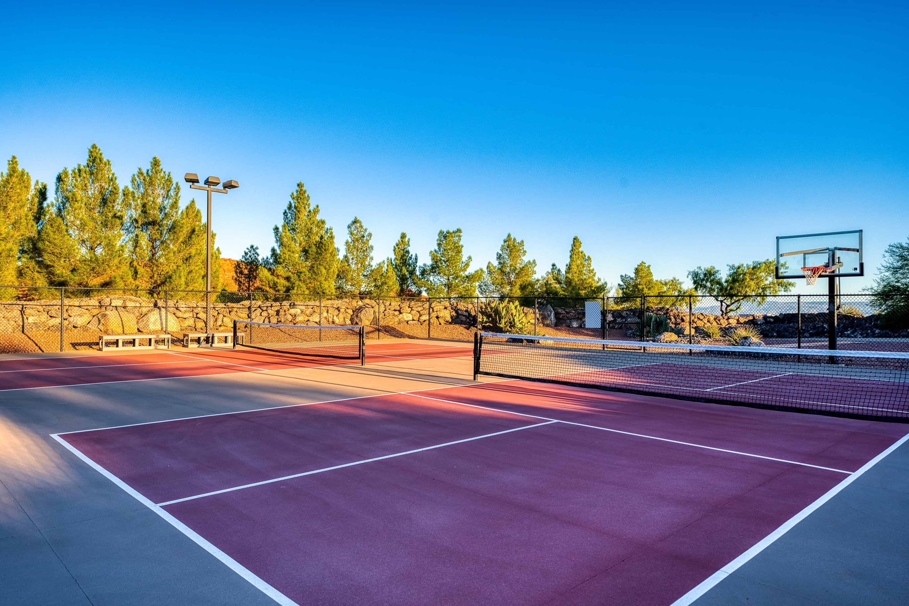 Cancha de tenis exterior con aro de baloncesto, rodeada de árboles y un muro de piedra.