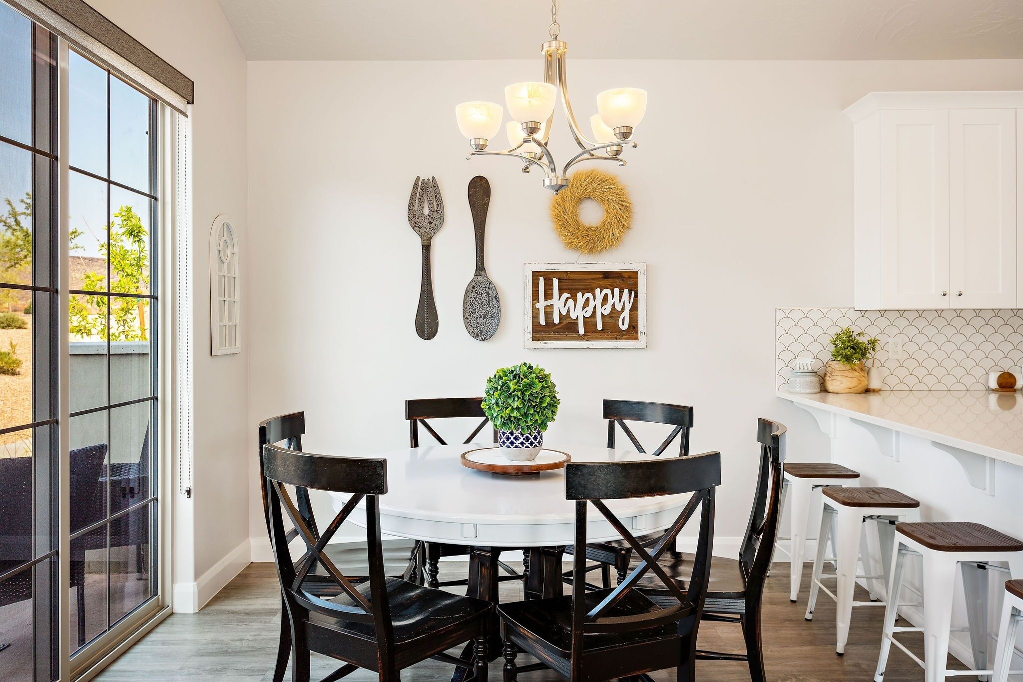 A dining area with a round table, black chairs, a potted plant, a chandelier, wall decorations, and a kitchen counter with stools.