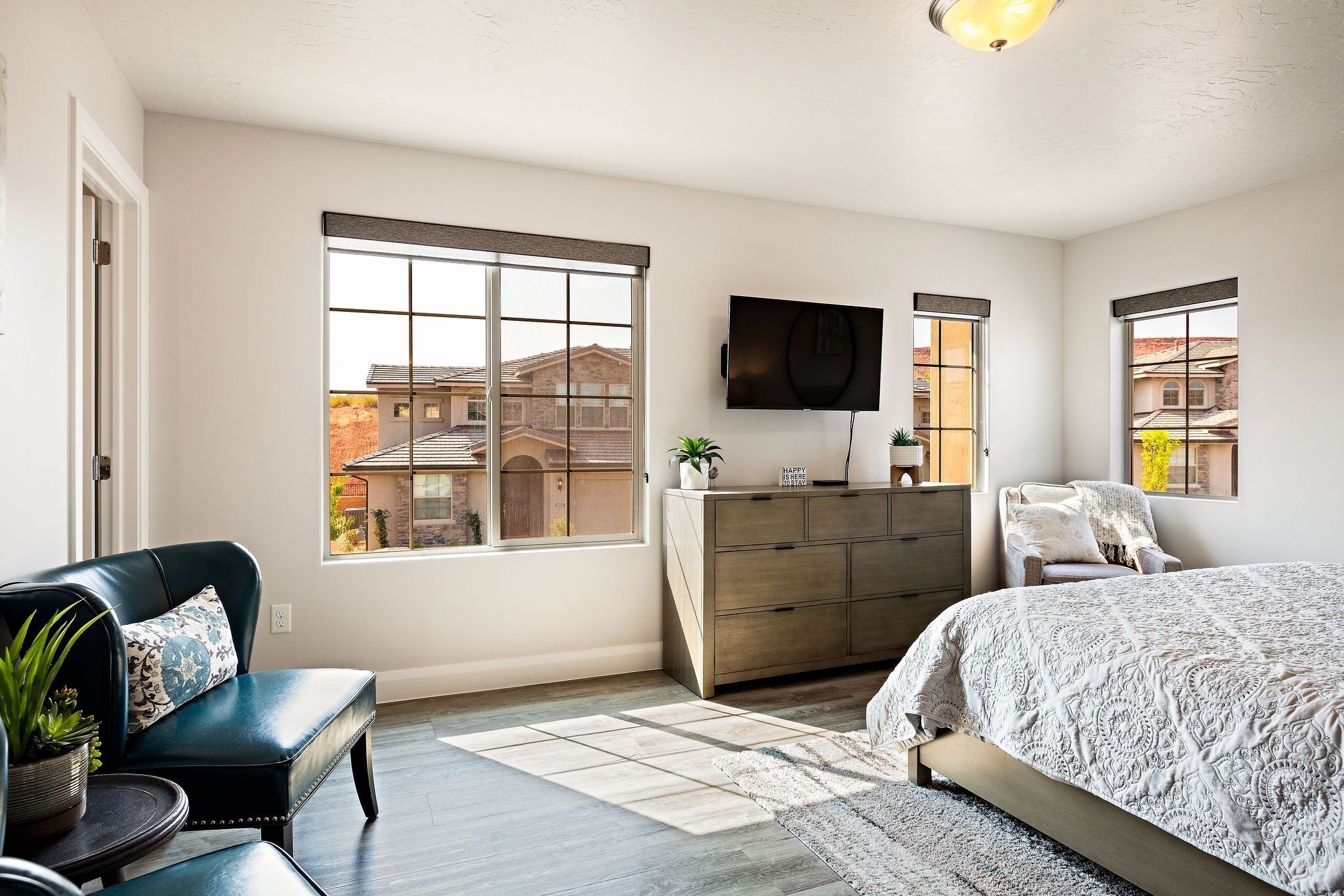 A bedroom with a large bed, a dresser, a TV, and two windows with a view of buildings.