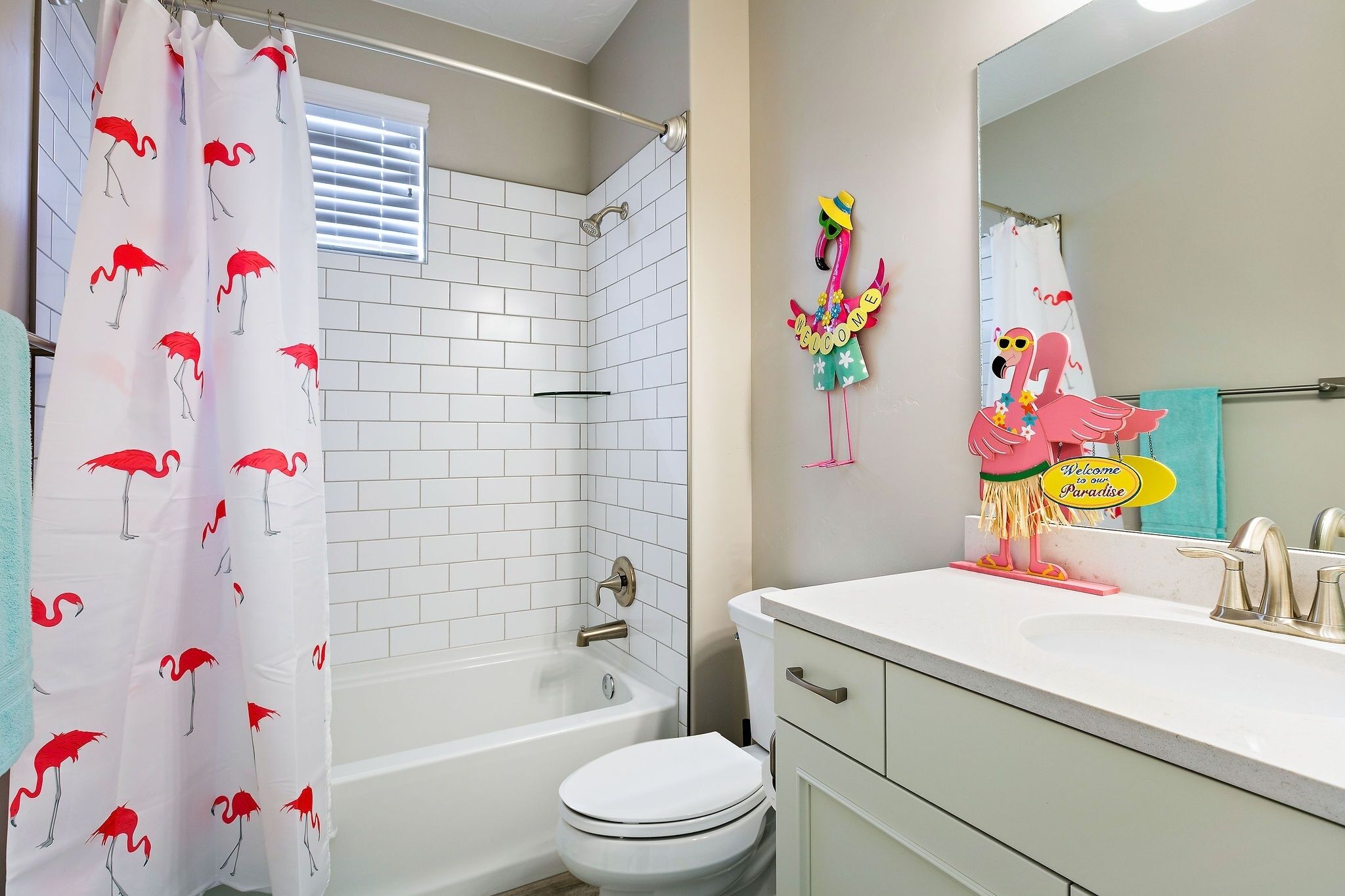 Une salle de bain avec une baignoire blanche, des toilettes et un lavabo. Il y a un rideau de douche orné de motifs de flamants roses, ainsi qu’un miroir décoré de flamants roses.