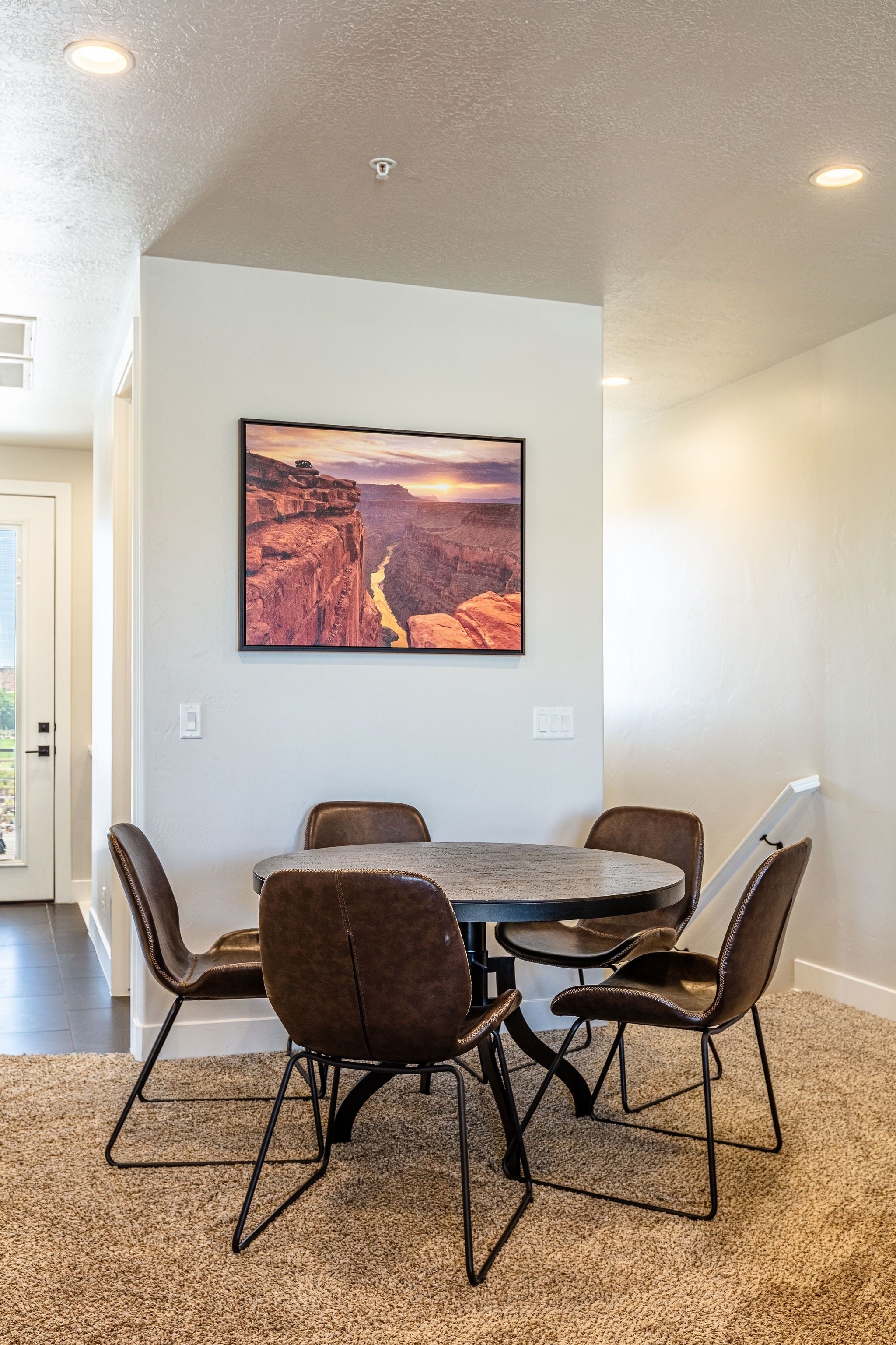 A dining area with a round table and six chairs, a large painting of a canyon on the wall, and a carpeted floor.