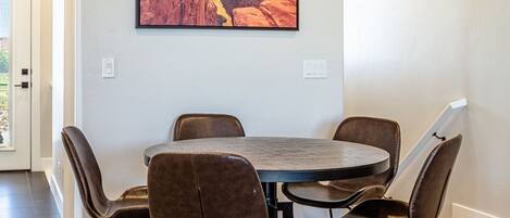 A dining area with a round table and six chairs, a large painting of a canyon on the wall, and a carpeted floor.
