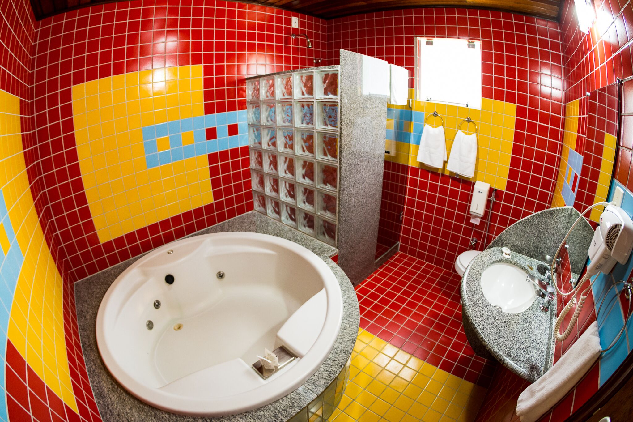 Casa de banho com parede revestida a azulejos vermelhos e amarelos, uma banheira branca, uma pia com espelho e um porta-toalhas.