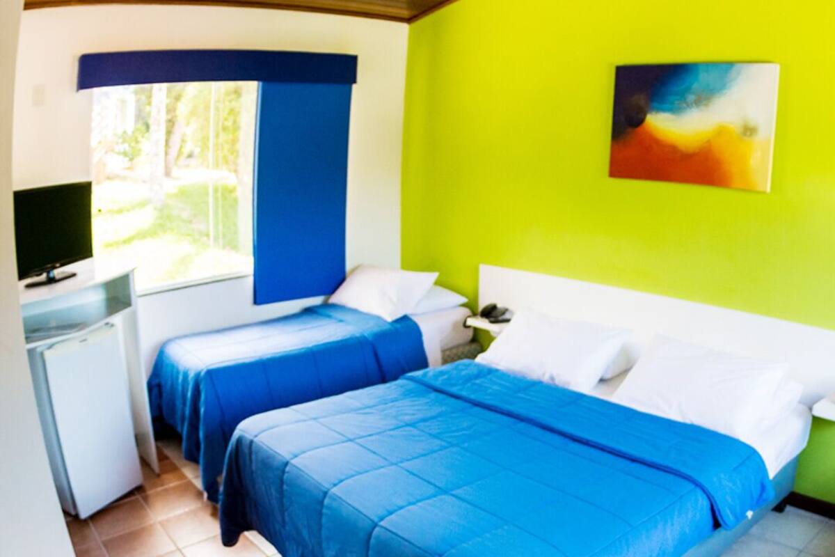 Una habitación con dos camas, un escritorio pequeño con una computadora y una ventana con cortinas azules.