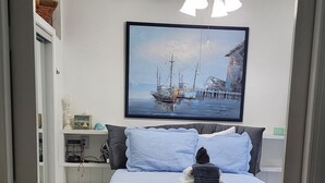 Une chambre à coucher avec un lit, un ventilateur de plafond, un tableau représentant un bateau et une petite étagère avec quelques objets.