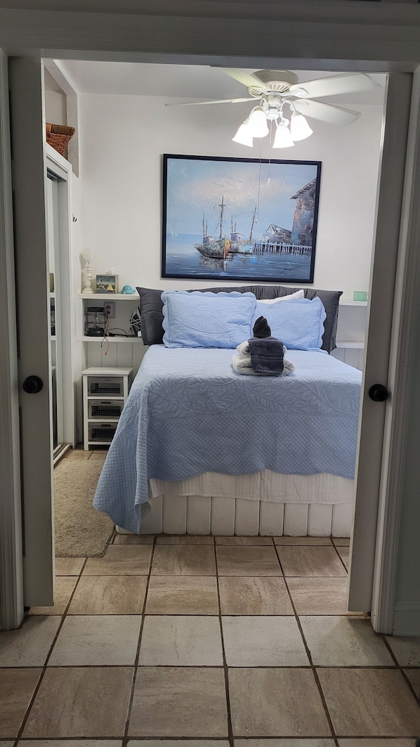 A bedroom with a bed, a ceiling fan, a painting of a boat, and a small shelf with items. - Minutes from the beach, with indoor hot tub (Daufuskie Island)